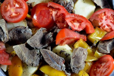 A tray of potato slices, onion, tomato, colored pepper, pieces of meat, spices, garlic and paprika, selective focus of potatoes with beef meat and vegetables ready to be baked The Egyptian way