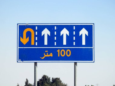 Arapça metnin çevirisi (100 M 100 metre) 4 şeridi gösteren ve sürücüleri sol şeritte 100 metre ileriden U dönüşü için yönlendiren bir yan trafik levhasında