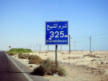 Sharm El Sheikh otobanındaki bir yol tabelası İngilizce yazılmış Sharm 325 km 'ye kalan mesafeyi ve caddenin kenarında Arapça ve İngilizce yazılmış Sharm Al Shiekh yazısını verir.