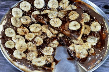 Fırında pişmiş, üzerinde erimiş çikolata ve taze muz ve susamlı dilimler olan tatlı bir çikolatalı pizza.