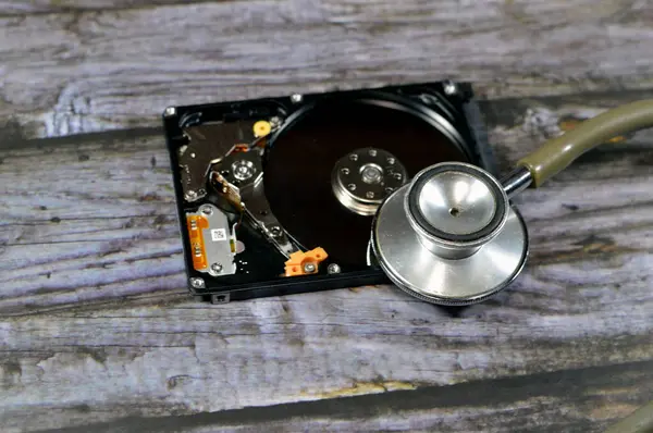 A medical stethoscope on a laptop 2.5 inch hard disk drive storage memory, repair broken computer part, an open hard disc with platters, spindle, actuator and read, write head, HDD diagnostic tool
