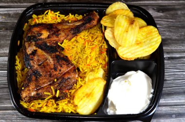 Arabic Syrian cuisine of charcoal grilled barbecued chicken with colorful Basmati rice and fried chips potatoes, served in a black disposable plate, served with regular and spicy garlic sauce