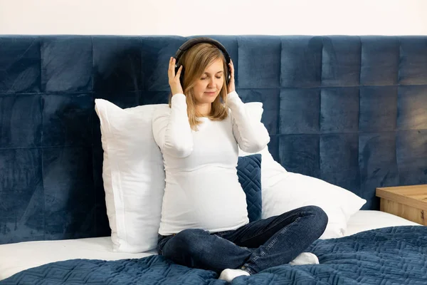 Beautiful pregnant woman with big belly wears headphones, sitting on bed, prenatal hypnosis, music listening, meditates. Baby delivery preparation. Happy childbearing period, caring for unborn baby