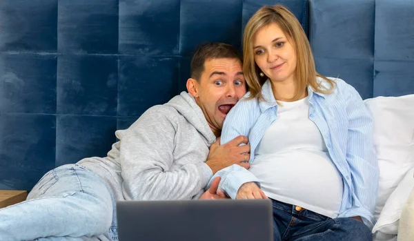 White Caucasian scared man, pregnant woman using, watching computer, laptop, sitting, lying on bed. Family, couple is preparing to baby delivery. Male wears in blue jeans and hoodie, female in shirt