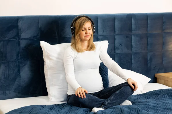Beautiful pregnant woman with big belly wears headphones,listens to hypnosis, music,meditates,sitting on bed. Baby delivery preparation. Happy childbearing period, caring for unborn baby. Female wears
