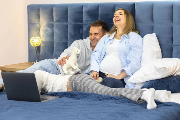 Happy family lying on bed watching video on notebook computer. Father plays with female child with lama toy, laughing pregnant mom. Cozy evening, spending time together. Horizontal. Casual clothes
