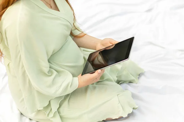 Cropped white pregnant girl holding, reading something on tablet,gadget,resting in park,sitting on cover,blanket in park,meadow. Healthy walk outdoors. Preparation for childbirth. Horizontal