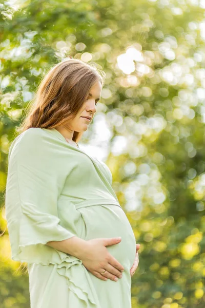 Young pregnant woman holds big belly, green trees,sky on background. Brown long haired female wear dress. Magic happy pregnancy. Child delivery preparations, emotional connection with baby. Vertical