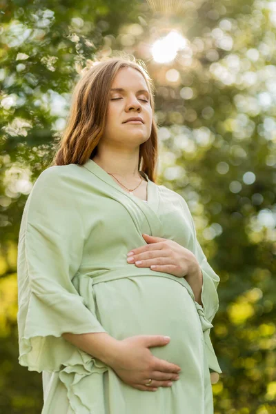 年轻的怀孕女性手挽着大肚子 闭着眼睛 背景上的阳光 快乐的女人穿裙子 神奇的怀孕劳动 与婴儿的情感联系 垂直平面 — 图库照片
