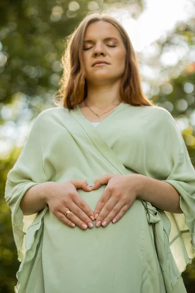 Happy pregnant woman makes love heart sign on her round belly. closed eyes, green trees, sunlight on background. White female wears dress.Magic pregnancy.Labor,emotional connection with baby.Vertical.