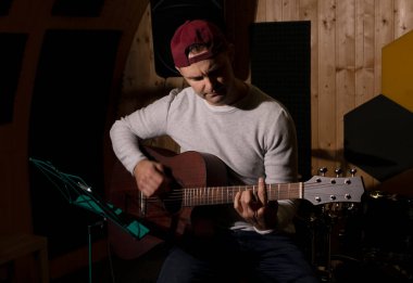 Beyaz adam gitar çalıyor, elinde müzik aleti akort ediyor, stüdyoda sandalyede oturuyor. Karanlık fotoğraf. Erkek, gündelik kıyafet giyer. Hobi, yaratıcı insanın boş zamanı, sanatçı. Uzayı kopyala Yatay