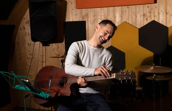 Smiling white man tuning guitar, playing, holding musical instrument in hands, sitting on chair in studio. Darken photo. Male wears casual cloth. Hobby, leisure of creative person, artist.Horizontal