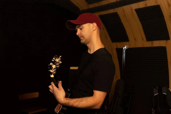 Caucasian man plays guitar, holding musical instrument in hands, standing in studio. Darken photo. Male wears casual cloth. Hobby, leisure of creative person, artist. Copy space. Horizontal