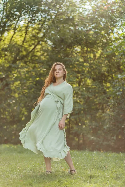 Dancing pretty regnant woman with big belly, green trees, meadow on background. White female wears dress. Magic happy pregnancy. Baby shower, childbirth, emotional connection with baby. Vertical plane