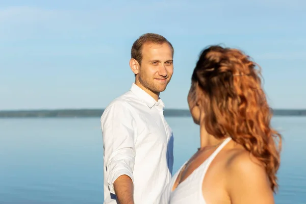 stock image Happy smiling Caucasian man holds hand of Latin woman, blue sky and water on background. International couple in love look at each other. Romantic sunny day. St Valentines day, enjoying togetherness