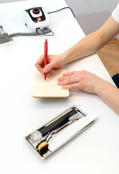 Unrecognizable nail master makes notes in notebook with pen, sitting at workstation, table in nail beauty treatment salon. Professional tools are on white table. Manicurists routine. Vertical plane