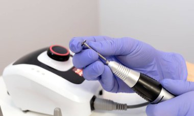 Manikürcü elektrikli donanım matkap tırnak makinesine nozül yerleştiriyor, mor koruluklar giyiyor. Kaplıca salonundaki manikür odası. Yakın plan, yatay. Manikür, pedikür konsepti. Yüksek kalite fotoğraf