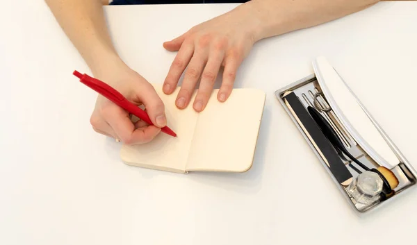 Unrecognizable nail master makes notes in notebook with pen, sitting at workstation, table in nail beauty treatment salon.Professional tools are on white table.Manicurists routine.Top view Horizontal