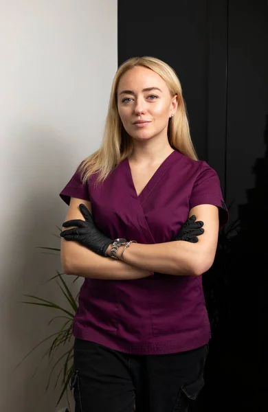 Beautiful female cosmetic physician, beauty doctor poses looking at camera, crossing hand on chests, wears medical scrubs, clothes on white black background. Cosmetology, aesthetic medicine. Vertical