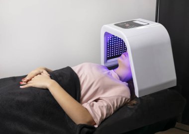 Yüz bakımı için LED ışık maskesi. Genç bir kadın, mavi maskenin altında kanepede uzanarak cilt tazeleme tedavisi görüyor. Kolajen üretiminin uyarılması, sivilce bakterilerinin öldürülmesi. Yatay
