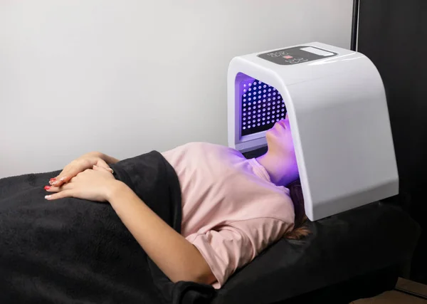 LED light mask for facial skin therapy, care. Young woman gets skin rejuvenation light treatment, lying on couch under blue mask. Stimulation of collagen production, killing acne bacteria. Horizontal
