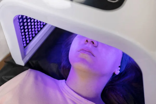 Blue led light mask for face skin therapy, care. Young girl gets problem skin treatment, lying on couch under blue light mask. Stimulation of collagen production, killing acne bacteria. Horizontal