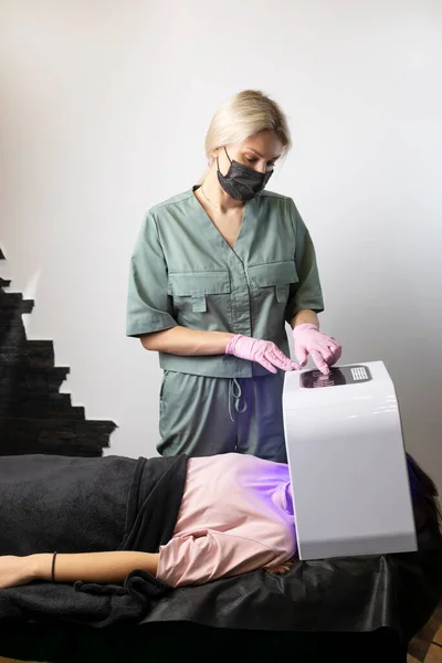 Beautician switches led light mask, device on. Young woman gets skin rejuvenation light treatment, lying on couch under blue mask. Facial skin therapy, care. Beauty face procedure. Vertical plane
