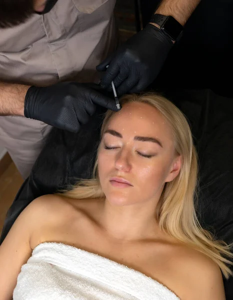 Erkek kozmetikçi, güzel bir kadının alnına, kırışıklık önleyici iğneler vuruyor. Güzellik salonunda kadın estetiği kozmetolojisi. Kozmetik konsepti, cilt bakımı. Dikey düzlem