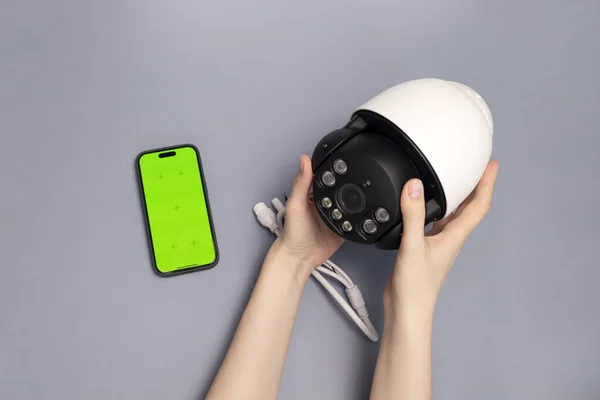 Human Hands Hold Surveillance IP Video Wireless Camera, Chroma Key Green Screen of Smartphone On Gray Background.Security Equipment For Smart Home with Online Remote App Monitoring, Control.Horizontal