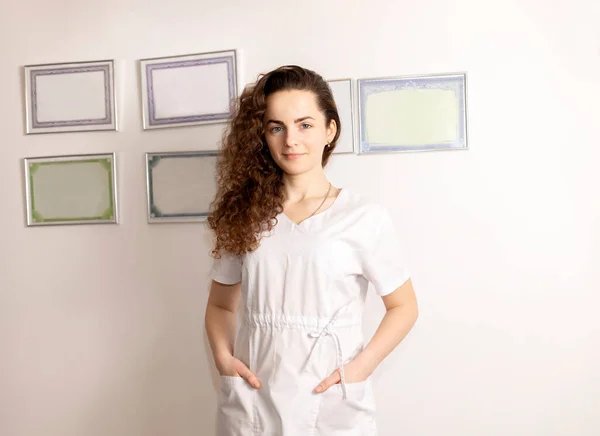 Portrait Smiling Young Female Rehabilitation Specialist, Physiotherapist In White Medical Clothes, Certificates In Frames On Wall On Background. Health Specialist, Rehabilitation. Horizontal plane.