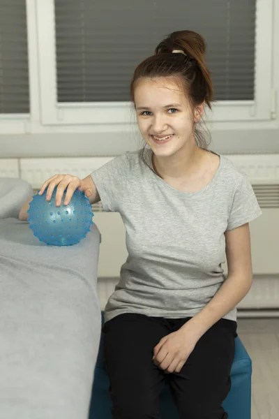 Portrait Of Happy Smiling Girl With Cerebral Palsy Sitting in Chair, Holding Blue Massage Ball with Pimples in Hospital. Rehabilitation. Balance And Posture Maintenance, Cure. Vertical plane.