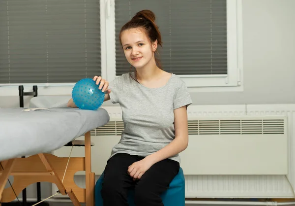 Portrait Of Cheerful Girl With Cerebral Palsy Sitting On Chair, Holding Blue Massage Ball With Pimples in Hospital. Rehabilitation. Balance And Posture Maintenance, Cure. Horizontal plane