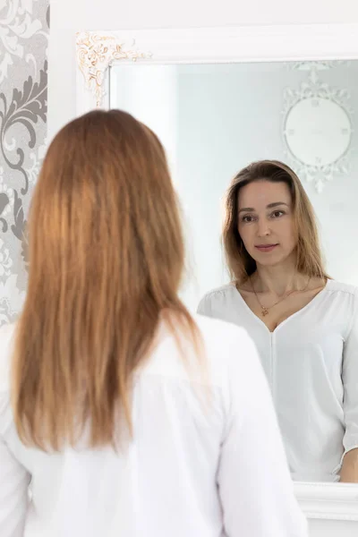 Çekici, esmer, beyaz tenli 40 Yo Kadını 'nın portresi aynaya bakıyor ve gülümsüyor. Sağlıklı Nemlendirilmiş Cilt Bakımı, Yaşlanan Güzellik, Cilt Tedavisi Kozmetik Konsepti. Dikey Düzlem