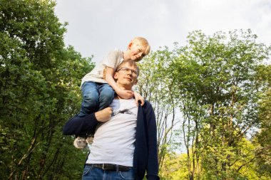 Baba, parkta yürürken küçük bir çocuğu omzunda taşıdı. Yaz zamanı. Mutlu Ebeveynlikler, Aile Boş Zamanları. Duygusal Bağlantı, Sevgi ve Bakım. Yatay düzlem. Yüksek kalite fotoğraf
