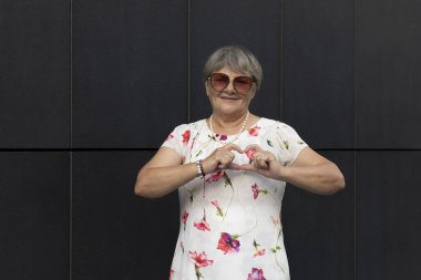 70 'li yaşlarda çok tatlı bir yaşlı kadın, elleriyle ve parmaklarıyla kalp şeklini gösteriyor, arkaplandaki karanlık duvarı. Gülümseyen yaşlı bir kadının portresi. Otantik Yaşam Tarzı Yüksek Kaliteli Fotoğraf