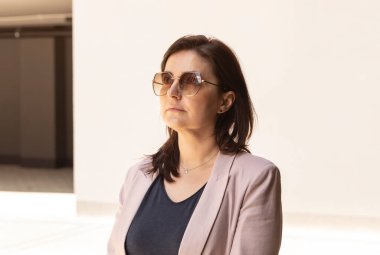 Kendine güvenen 40 'lı yaşlardaki güzel kadının portresi. Güneş Gözlüklü Beyaz Burnette Kadın Uzaklara Bakar, Pembe Ceket Giyer, Mavi Gömlekli. Yaz zamanı. Yatay Düzlem.