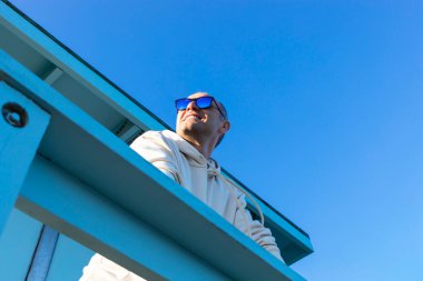 Gülümseyen Orta Çağ beyaz erkek tek başına tahta iskelede duruyor, veranda, arka planda mavi gökyüzü. Güneşli bir gün. Kapüşonlu kazak, kot pantolon, güneş gözlüğü takan bir adam. Uzayı kopyala Tatil Gevşemesi. Huzurlu.