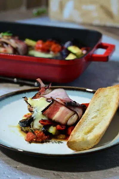 Kızarmış peynirli peynirin yakın plan çekimi. Çıtır pastırmaya sarılmış, taze otlarla süslenmiş ve yanında renkli ratatouille sebzeleriyle servis edilmiş.