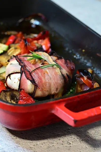 Kızarmış peynirli peynirin yakın plan çekimi. Çıtır pastırmaya sarılmış, taze otlarla süslenmiş ve yanında renkli ratatouille sebzeleriyle servis edilmiş.