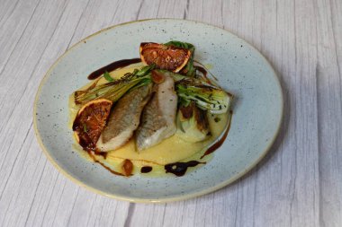 Koyu morina filetosu, kremalı polenta, kan portakalı segmentleri ve açık arkaplanda taze yeşil salata içeren güzel bir ev yapımı yemeğin üst görüntüsü.
