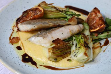 Koyu morina filetosu, kremalı polenta, kan portakalı segmentleri ve açık arkaplanda taze yeşil salata içeren güzel bir ev yapımı yemeğin üst görüntüsü.