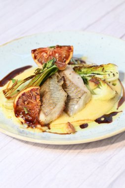 Koyu morina filetosu, kremalı polenta, kan portakalı segmentleri ve açık arkaplanda taze yeşil salata içeren güzel bir ev yapımı yemeğin üst görüntüsü.