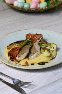 Koyu morina filetosu, kremalı polenta, kan portakalı segmentleri ve açık arkaplanda taze yeşil salata içeren güzel bir ev yapımı yemeğin üst görüntüsü.