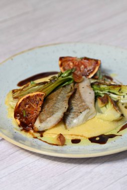 Koyu morina filetosu, kremalı polenta, kan portakalı segmentleri ve açık arkaplanda taze yeşil salata içeren güzel bir ev yapımı yemeğin üst görüntüsü.