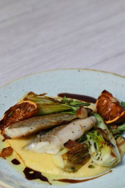Koyu morina filetosu, kremalı polenta, kan portakalı segmentleri ve açık arkaplanda taze yeşil salata içeren güzel bir ev yapımı yemeğin üst görüntüsü.
