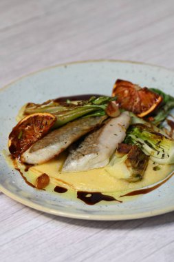 Koyu morina filetosu, kremalı polenta, kan portakalı segmentleri ve açık arkaplanda taze yeşil salata içeren güzel bir ev yapımı yemeğin üst görüntüsü.