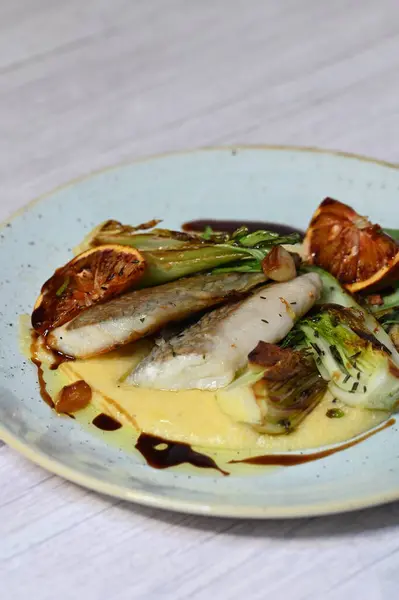 Koyu morina filetosu, kremalı polenta, kan portakalı segmentleri ve açık arkaplanda taze yeşil salata içeren güzel bir ev yapımı yemeğin üst görüntüsü.