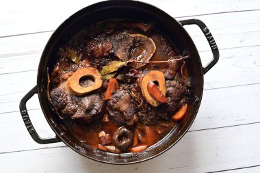 Kırmızı şarapta pişirilmiş kemikli yumuşak sığır eti, taze otlar ve tek bir kırsal siyah tencerede kök sebzelerle servis edilir..