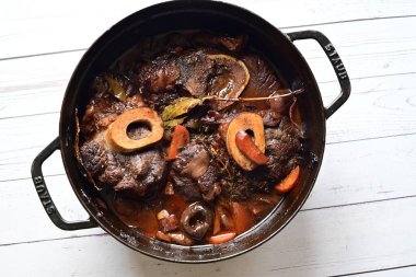 Kırmızı şarapta pişirilmiş kemikli yumuşak sığır eti, taze otlar ve tek bir kırsal siyah tencerede kök sebzelerle servis edilir..