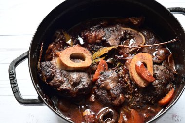 Kırmızı şarapta pişirilmiş kemikli yumuşak sığır eti, taze otlar ve tek bir kırsal siyah tencerede kök sebzelerle servis edilir..